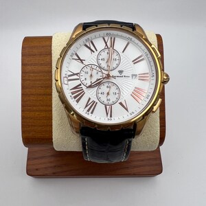 May include: A gold-toned Raymond Rene watch with a white face, featuring Roman numerals and three sub-dials. The watch has a black leather band and is displayed on a wooden stand. The watch face has rose gold accents.