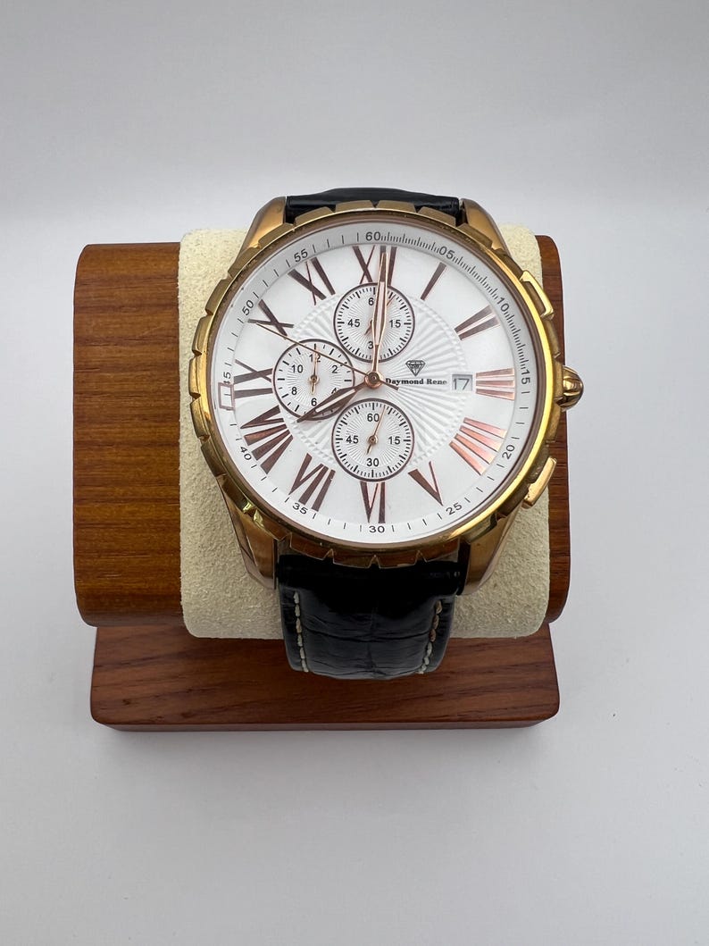 May include: A gold-toned Raymond Rene watch with a white face, featuring Roman numerals and three sub-dials. The watch has a black leather band and is displayed on a wooden stand. The watch face has rose gold accents.