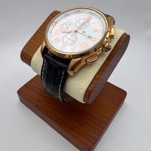 May include: A gold-toned chronograph watch with a white face, Roman numeral hour markers, and three sub-dials. The watch has a black leather band with white stitching and is displayed on a wooden watch stand.