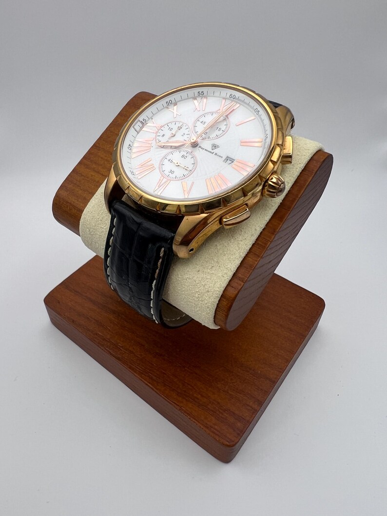 May include: A gold-toned chronograph watch with a white face, Roman numeral hour markers, and three sub-dials. The watch has a black leather band with white stitching and is displayed on a wooden watch stand.