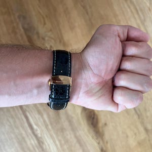 May include: A black leather watch band with white stitching and a gold-colored clasp is fastened on a wrist. The watch band has a crocodile skin texture. The watch band is attached to a gold-colored watch face.