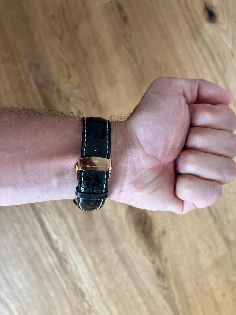 May include: A black leather watch band with white stitching and a gold-colored clasp is fastened on a wrist. The watch band has a crocodile skin texture. The watch band is attached to a gold-colored watch face.