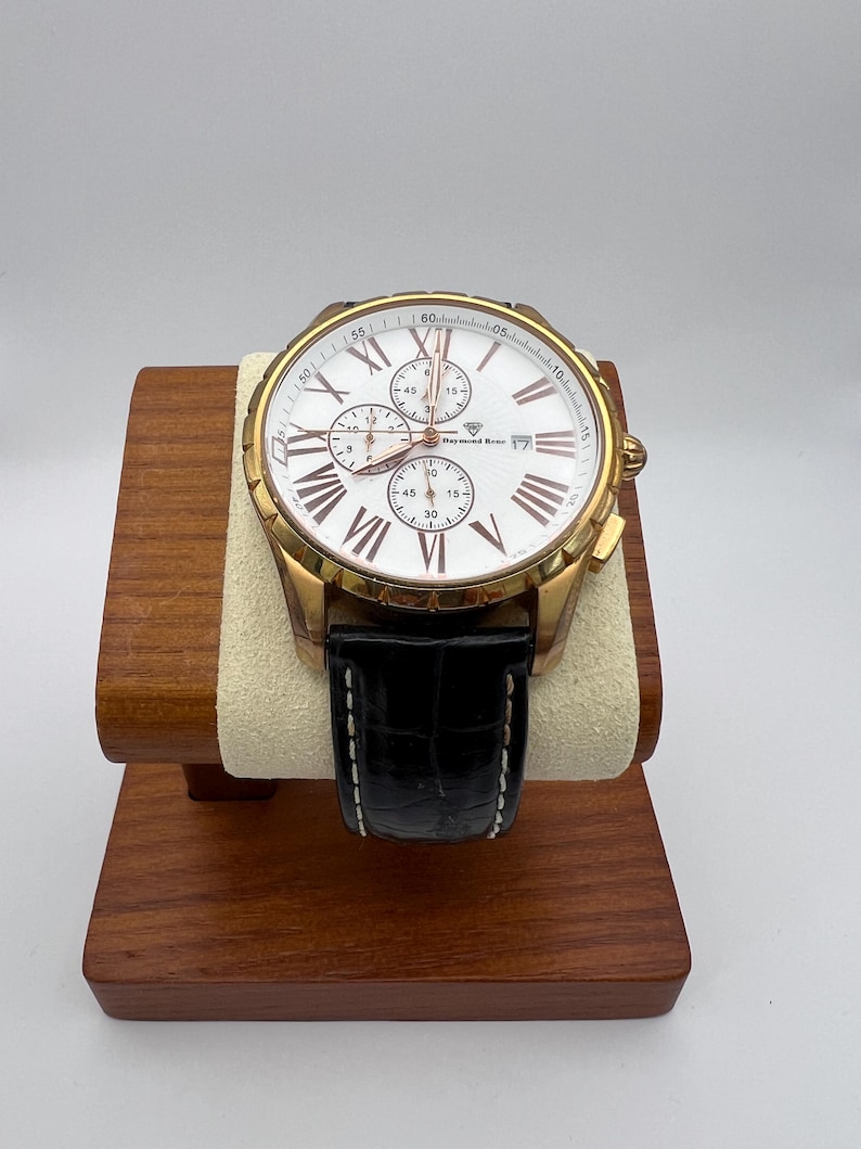 May include: A gold-toned wristwatch with a white face, Roman numeral hour markers, and three sub-dials. The watch has a black leather band and is displayed on a wooden stand. The watch face has the text "Raymond Weil".