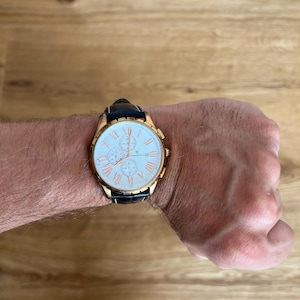 May include: A gold-toned wristwatch with a white face and black leather band. The watch features Roman numeral hour markers and three sub-dials. The watch is on a wrist.