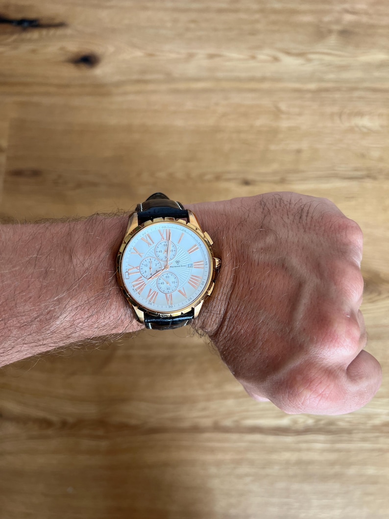 May include: A gold-toned wristwatch with a white face and black leather band. The watch features Roman numeral hour markers and three sub-dials. The watch is on a wrist.