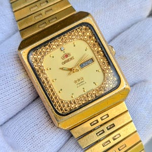 Puede incluir: Reloj de pulsera Orient dorado con esfera rectangular. La esfera es de color crema con agujas e indicadores dorados. El bisel está incrustado con pequeños cristales. La correa también es dorada.