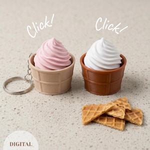 Puede incluir: Dos llaveros de cono de helado con partes superiores en espiral rosa y blanca, en copas de barquillo marrón claro y oscuro. El texto Click! aparece sobre cada uno, y DIGITAL PRODUCT está en un círculo. Varios trozos de barquillo rotos cerca.