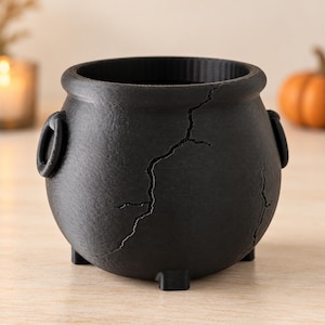 May include: A matte black cauldron-shaped container with a textured surface and a prominent vertical crack running down the front. The vessel features two side handles, a wide circular opening, and stands on three small, short feet.
