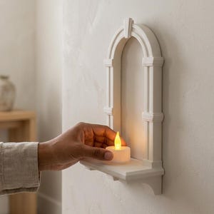 Könnte beinhalten: Ein weißes, bogenförmiges Wandregal mit architektonischen Details und einer kleinen Ablagefläche. Ein batteriebetriebenes Teelicht mit einer warmgelben Flamme steht auf dem Regal und wirft einen sanften Schein auf die Struktur und die Wand.