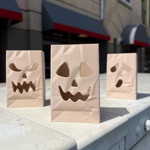 May include: Three light beige, rectangular, upright paper-like bags featuring die-cut jack-o'-lantern faces. Each bag displays a unique facial expression with cutouts for eyes, noses, and jagged mouths, mimicking the appearance of creased paper.
