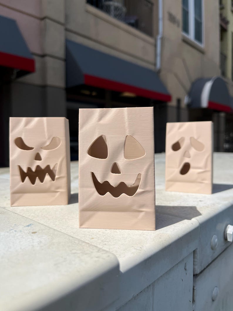 May include: Three light beige, rectangular, upright paper-like bags featuring die-cut jack-o'-lantern faces. Each bag displays a unique facial expression with cutouts for eyes, noses, and jagged mouths, mimicking the appearance of creased paper.
