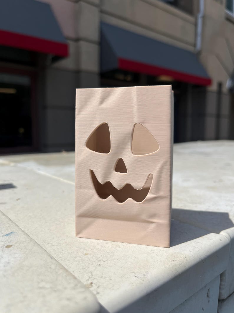 May include: A light beige, rectangular paper bag featuring a die-cut jack-o'-lantern face. The design includes two triangular eye cutouts, a small triangular nose cutout, and a wide, wavy-edged mouth cutout, creating a hollow facial expression.