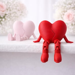 Op de afbeelding: Twee hartvormige beeldjes, één roze en één rood, elk met een scheur in het midden. Het rode hart heeft beweegbare benen en armen en lijkt op een wit oppervlak te zitten. Achtergrond met zacht gefocuste witte en roze bloemen.