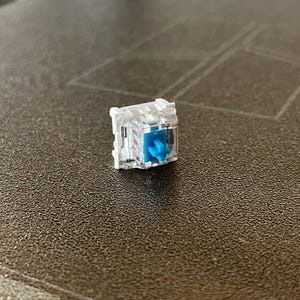 K&ouml;nnte beinhalten: Ein mechanischer Tastaturschalter mit einem transparenten Kunststoffgeh&auml;use und einem hellblauen, kreuzf&ouml;rmigen Schaft. Die internen Komponenten sind durch das klare Geh&auml;use sichtbar und zeigen den Schaltmechanismus sowie den zentralen blauen Schaft.