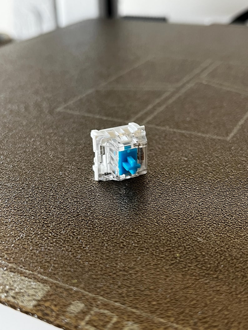 K&ouml;nnte beinhalten: Ein mechanischer Tastaturschalter mit einem transparenten Kunststoffgeh&auml;use und einem hellblauen, kreuzf&ouml;rmigen Schaft. Die internen Komponenten sind durch das klare Geh&auml;use sichtbar und zeigen den Schaltmechanismus sowie den zentralen blauen Schaft.
