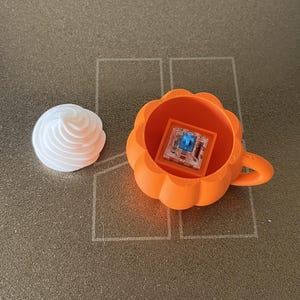K&ouml;nnte beinhalten: Ein leuchtend orangefarbener, k&uuml;rbisf&ouml;rmiger Beh&auml;lter mit Griff, der einen transparenten mechanischen Tastaturschalter mit blauem Stift enth&auml;lt, begleitet von einem separaten wei&szlig;en, wirbelf&ouml;rmigen Aufsatz.