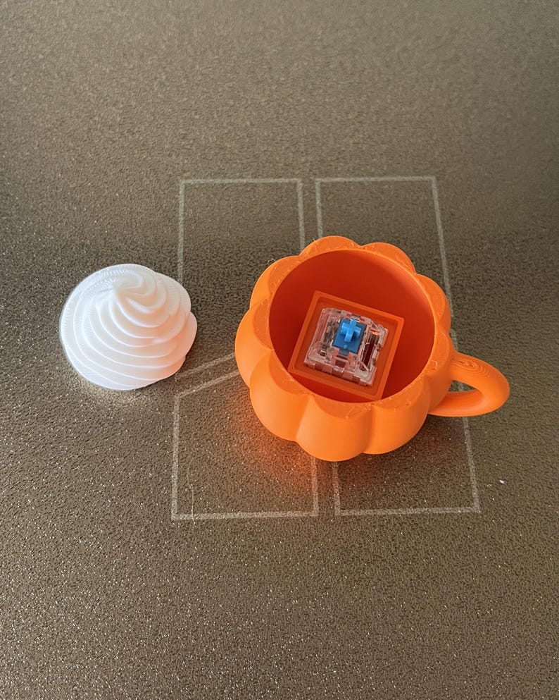 K&ouml;nnte beinhalten: Ein leuchtend orangefarbener, k&uuml;rbisf&ouml;rmiger Beh&auml;lter mit Griff, der einen transparenten mechanischen Tastaturschalter mit blauem Stift enth&auml;lt, begleitet von einem separaten wei&szlig;en, wirbelf&ouml;rmigen Aufsatz.