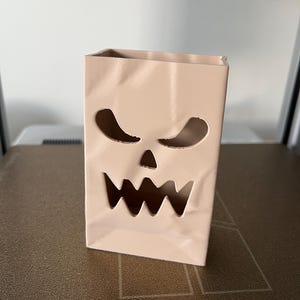 May include: A light beige, rectangular, upright container with a hollowed-out, jagged, scary face design featuring two triangular eyes and a wide, toothy mouth. The surface has subtle, molded creases that mimic the appearance of a folded paper bag.