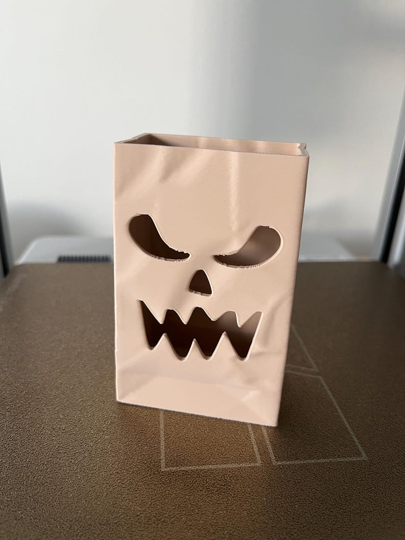 May include: A light beige, rectangular, upright container with a hollowed-out, jagged, scary face design featuring two triangular eyes and a wide, toothy mouth. The surface has subtle, molded creases that mimic the appearance of a folded paper bag.