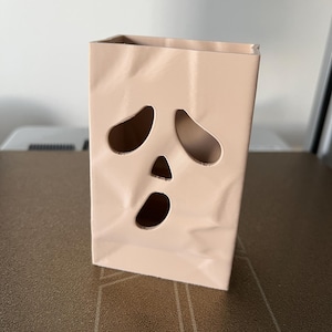 May include: A light beige, rectangular, hollow container with a textured, crumpled paper-like surface. The front features three cut-out openings arranged to resemble a stylized, surprised face with two large, teardrop-shaped eyes and a small, oval mouth.