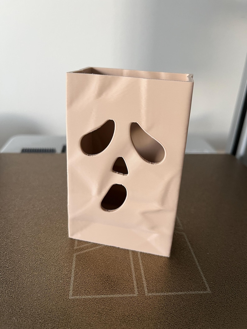 May include: A light beige, rectangular, hollow container with a textured, crumpled paper-like surface. The front features three cut-out openings arranged to resemble a stylized, surprised face with two large, teardrop-shaped eyes and a small, oval mouth.
