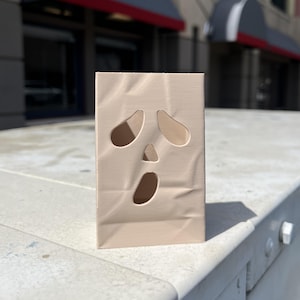 May include: A light beige, rectangular, 3D-printed object with a hollow interior and a ghost-like face design. The front surface features two oval-shaped eye cutouts, a small triangular nose cutout, and a large oval mouth cutout, mimicking a classic ghost expression.