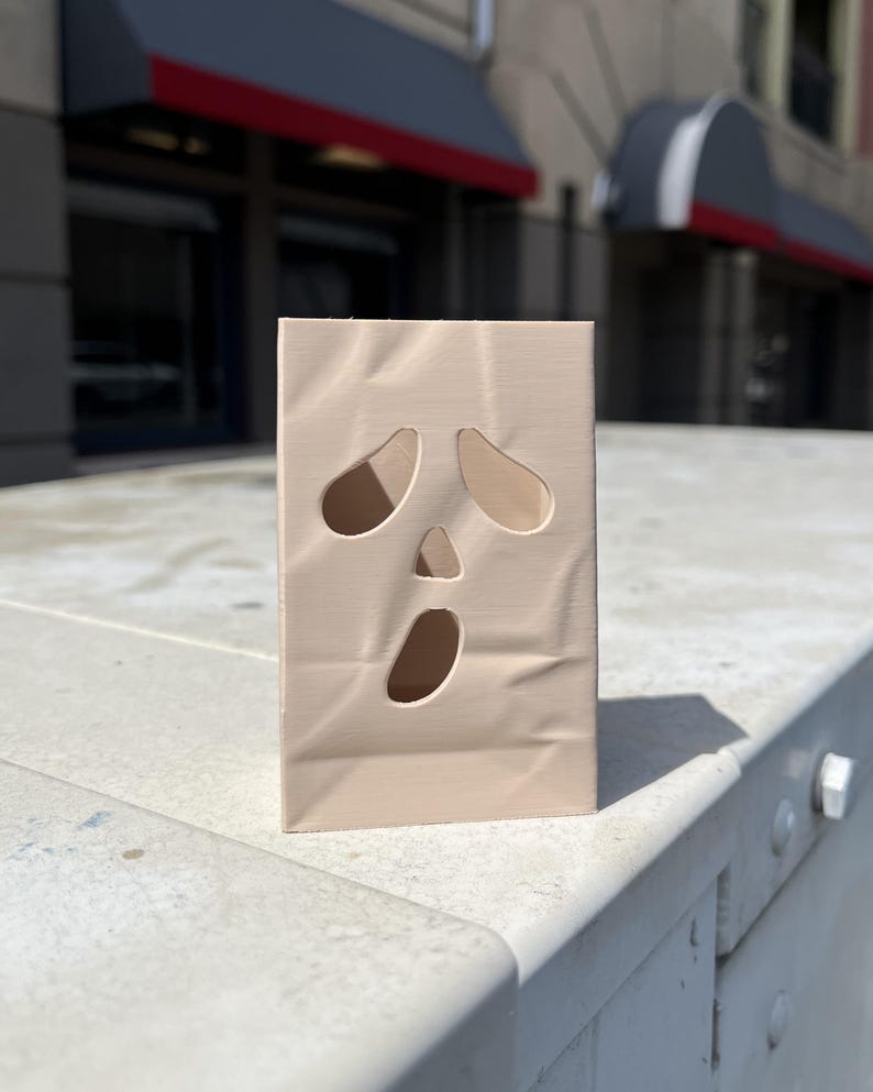 May include: A light beige, rectangular, 3D-printed object with a hollow interior and a ghost-like face design. The front surface features two oval-shaped eye cutouts, a small triangular nose cutout, and a large oval mouth cutout, mimicking a classic ghost expression.