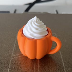 K&ouml;nnte beinhalten: Ein 3D-gedruckter, orangefarbener Tassenbecher in K&uuml;rbisform mit Henkel, der mit einem wei&szlig;en, spiralf&ouml;rmigen Aufsatz versehen ist, der an Schlagsahne erinnert. Das Objekt hat eine matte Textur und besteht aus Kunststoff mit sichtbaren Drucklinien.