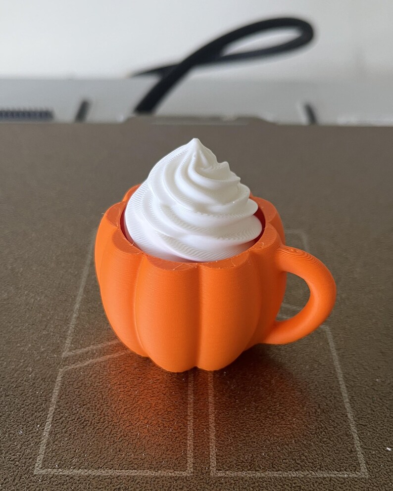 K&ouml;nnte beinhalten: Ein 3D-gedruckter, orangefarbener Tassenbecher in K&uuml;rbisform mit Henkel, der mit einem wei&szlig;en, spiralf&ouml;rmigen Aufsatz versehen ist, der an Schlagsahne erinnert. Das Objekt hat eine matte Textur und besteht aus Kunststoff mit sichtbaren Drucklinien.