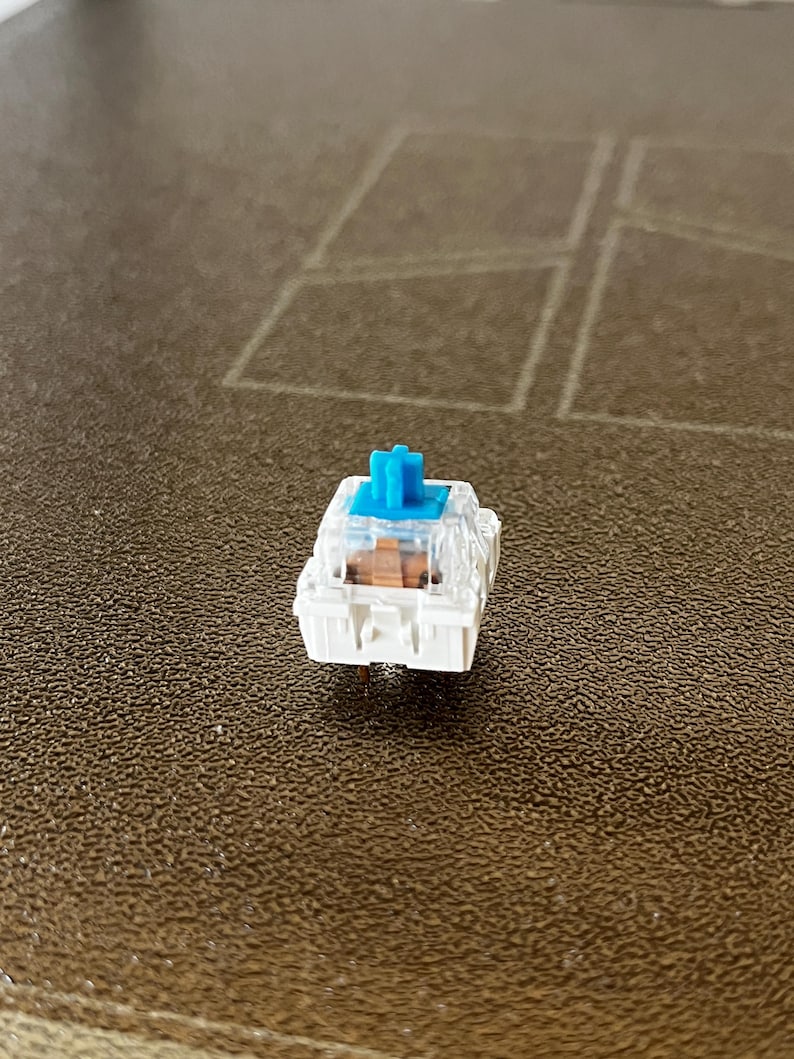 K&ouml;nnte beinhalten: Ein mechanischer Tastaturschalter mit wei&szlig;er Basis, transparentem Geh&auml;use und einem leuchtend blauen Schaft. Die internen Komponenten umfassen einen braunen Schieber, der durch das transparente Geh&auml;use sichtbar ist.