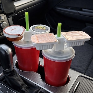 May include: A car cup holder with two red cups, each with a white lid and green straw. The holder also has spaces for sauce containers. The BBQ sauce container is brown, and the other containers are white and clear.