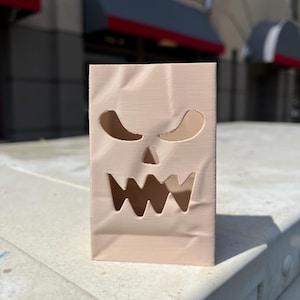 May include: A light beige, rectangular, upright object with a textured surface resembling a paper bag. It features cut-out shapes forming a stylized, jagged-toothed face with two triangular eyes and a wide, serrated mouth opening.