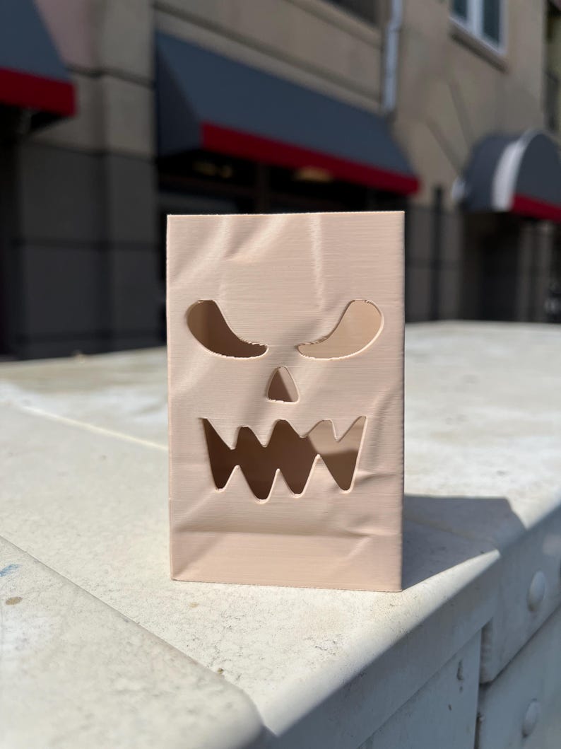 May include: A light beige, rectangular, upright object with a textured surface resembling a paper bag. It features cut-out shapes forming a stylized, jagged-toothed face with two triangular eyes and a wide, serrated mouth opening.