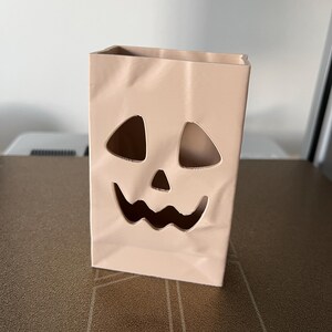 May include: A light beige, rectangular, paper-bag-style container featuring a cutout jack-o'-lantern face with two triangular eyes, a small triangular nose, and a wide, jagged, smiling mouth. The surface has subtle, molded, vertical creases.