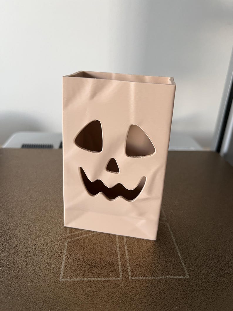May include: A light beige, rectangular, paper-bag-style container featuring a cutout jack-o'-lantern face with two triangular eyes, a small triangular nose, and a wide, jagged, smiling mouth. The surface has subtle, molded, vertical creases.