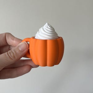 K&ouml;nnte beinhalten: Eine kleine orangefarbene Tasse in K&uuml;rbisform mit einem Henkel auf der linken Seite. Die Tasse ist mit einem wei&szlig;en, spiralf&ouml;rmigen Belag gef&uuml;llt, der an Schlagsahne erinnert und &uuml;ber den Rand des k&uuml;rbisf&ouml;rmigen Gef&auml;&szlig;es hinausragt.