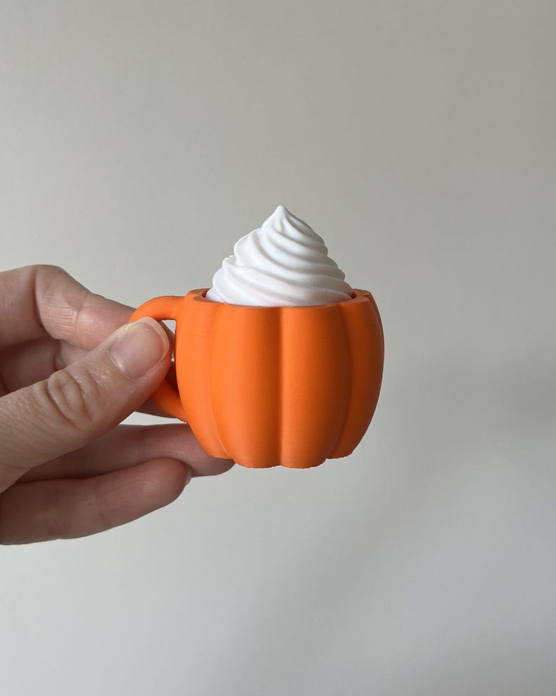 K&ouml;nnte beinhalten: Eine kleine orangefarbene Tasse in K&uuml;rbisform mit einem Henkel auf der linken Seite. Die Tasse ist mit einem wei&szlig;en, spiralf&ouml;rmigen Belag gef&uuml;llt, der an Schlagsahne erinnert und &uuml;ber den Rand des k&uuml;rbisf&ouml;rmigen Gef&auml;&szlig;es hinausragt.