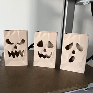May include: Three light beige, rectangular, upright containers with a crinkled paper bag texture. Each container features a unique, hollowed-out jack-o'-lantern face with cutouts for eyes, noses, and mouths, creating a spooky, decorative aesthetic.