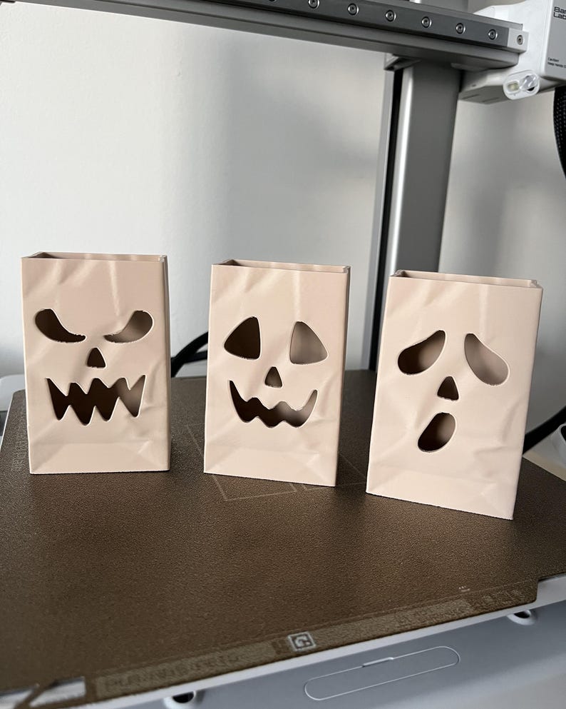 May include: Three light beige, rectangular, upright containers with a crinkled paper bag texture. Each container features a unique, hollowed-out jack-o'-lantern face with cutouts for eyes, noses, and mouths, creating a spooky, decorative aesthetic.