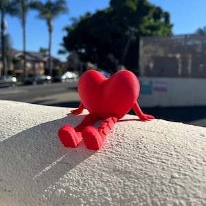 Puede incluir: Una figura roja brillante en forma de corazón con brazos y piernas, sentada sobre una superficie blanca texturizada. Las piernas de la figura están dobladas, como si estuviera relajándose. El fondo presenta palmeras y un cielo azul claro.