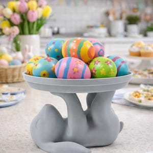 Op de afbeelding: Een grijs konijnvormig serveerblad toont een verzameling kleurrijke, versierde paaseieren. De eieren zijn beschilderd met verschillende ontwerpen, waaronder strepen, bloemen en stippen. Een boeket tulpen en andere paaslekkernijen staan op de achtergrond.