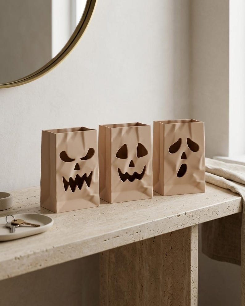 May include: Three light beige ceramic containers shaped like crumpled paper bags, each featuring a unique cutout face design. The first has a jagged grin, the second a triangular-eyed smile, and the third a wide-mouthed, surprised expression.