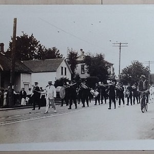 May include: Black and white photograph of a marching band parading down a street. The band members are in uniform, playing instruments. Spectators line the sidewalks. Two people ride bicycles in the street. Houses and trees are in the background.