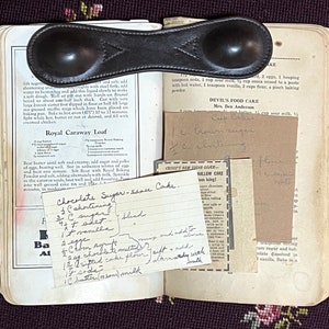 May include: Open vintage cookbook with handwritten recipes, including "Chocolate Sugar-sance Cake" and "Devil's Food Cake." A leather measuring spoon rests on the pages. The book is open on a floral patterned surface.