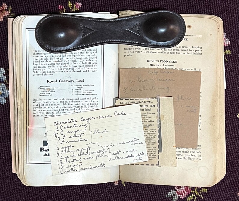 May include: Open vintage cookbook with handwritten recipes, including "Chocolate Sugar-sance Cake" and "Devil's Food Cake." A leather measuring spoon rests on the pages. The book is open on a floral patterned surface.