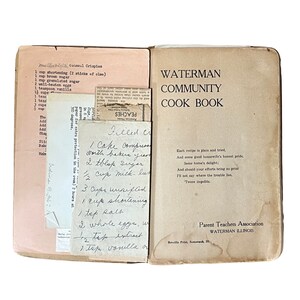 May include: An open vintage cookbook titled "Waterman Community Cook Book" with handwritten recipes and a pink recipe card. The cookbook is tan with black text and a Parent Teachers Association logo. The recipes include "Oatmeal Crispies" and "Filled Cream".
