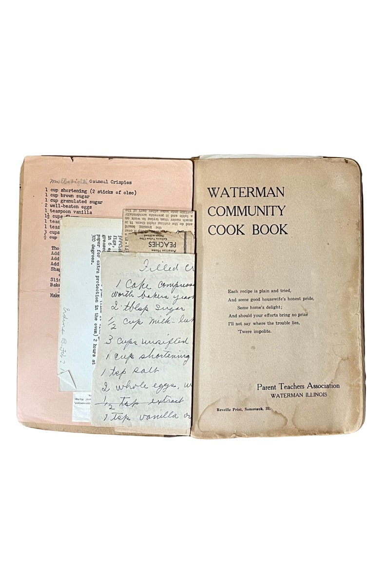 May include: An open vintage cookbook titled "Waterman Community Cook Book" with handwritten recipes and a pink recipe card. The cookbook is tan with black text and a Parent Teachers Association logo. The recipes include "Oatmeal Crispies" and "Filled Cream".