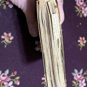 May include: A vintage cookbook with aged, yellowed pages. A small paper insert with text is visible. The book is held by a hand, with a floral patterned background. The text on the insert reads "minute frosting is one of..."