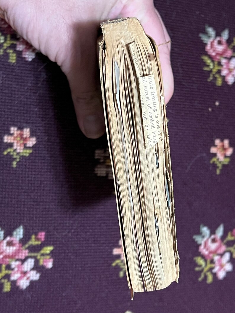 May include: A vintage cookbook with aged, yellowed pages. A small paper insert with text is visible. The book is held by a hand, with a floral patterned background. The text on the insert reads "minute frosting is one of..."