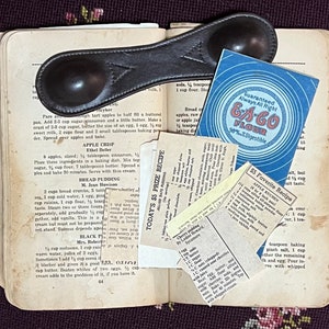 May include: An open vintage cookbook displays handwritten recipes and a leather measuring spoon. Recipe cards and a blue and white E-A-CO Flour advertisement are tucked inside. The theme is baking and cooking.